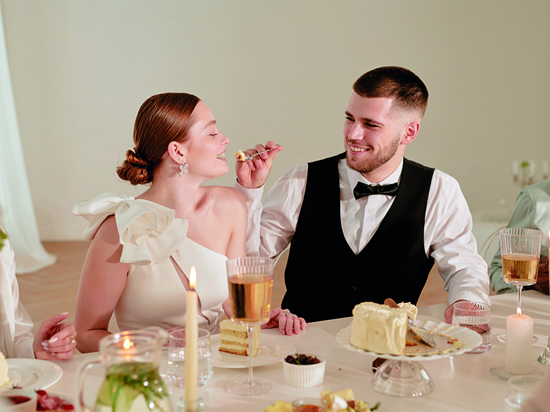Young Caucasian Bride and Groom Enjoying Wedding Reception Celebration Together Caucasian young adult man feeding cake to Caucasian young adult woman during wedding celebration, both smiling and sitting at decorated table with desserts and drinks, other guests partially visible