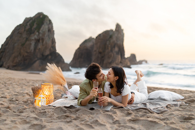 Happy couple having romantic date picnic on the beach, lying on blanket and drinking wine, having tender moment Happy couple having romantic date picnic on the beach near ocean, lying on blanket and drinking wine, having tender moment and enjoying time together, copy space