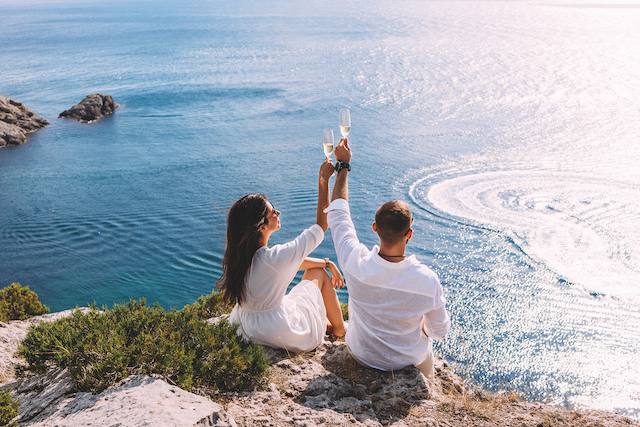 A,Couple,In,Love,Celebrates,Their,Engagement,On,The,Seashore. A couple in love celebrates their engagement on the seashore. A beautiful couple drinks champagne by the sea. Honeymoon trip. Lovers on the beach. Wedding travel. Couple on vacation. Copy space
